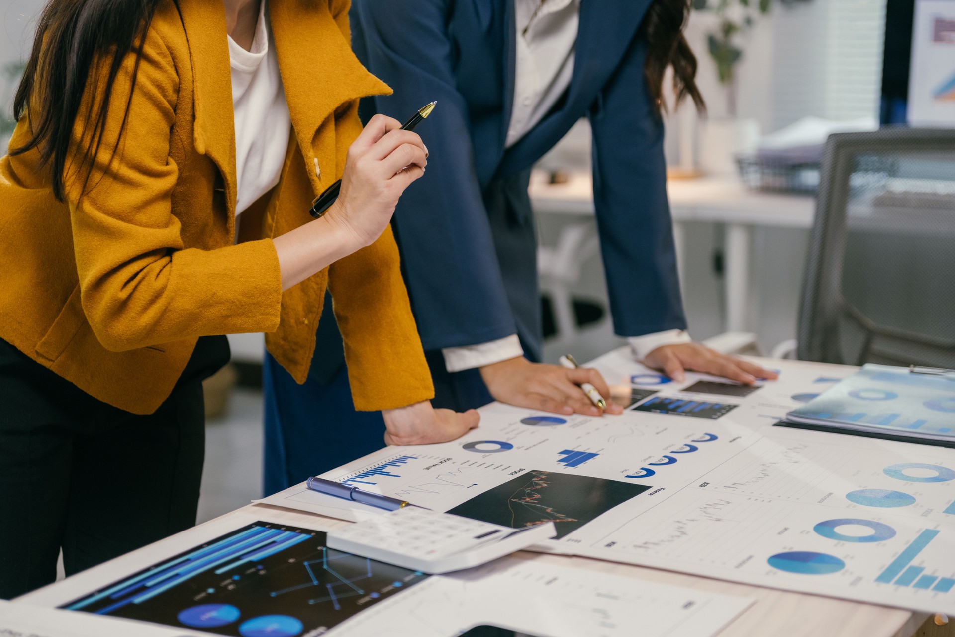 Two businesswomen are examining financial charts and reports, discussing data analysis and strategic planning in a modern office setting